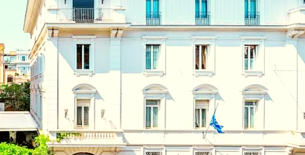Elegant white hotel facade with arched windows, blue sky, cobblestone street, green plants, and "Hotel Locarno" text. Calm, sunny atmosphere.