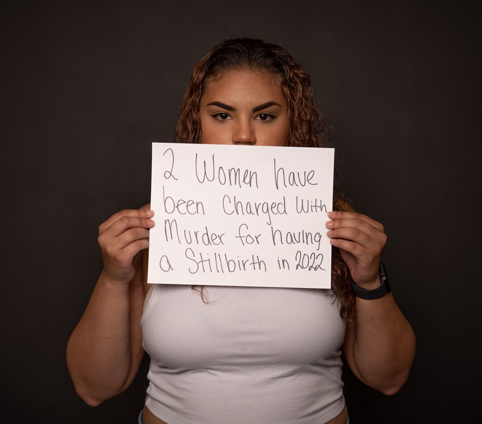 Nayeliz holding a sign partially covering her face that says "2 Women have been charged with murder for having a stillbirth in 2022." 