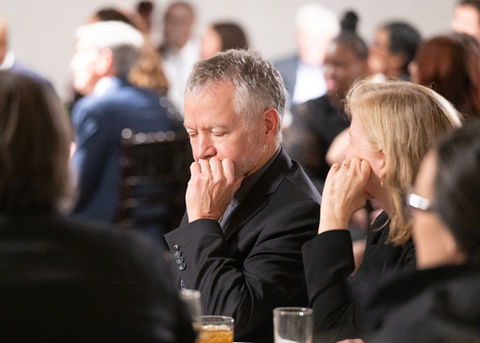 A man dressed in a suit at a gala sits at a table, resting his chin on his hand, deep in thought or contemplation listening to the speaker.