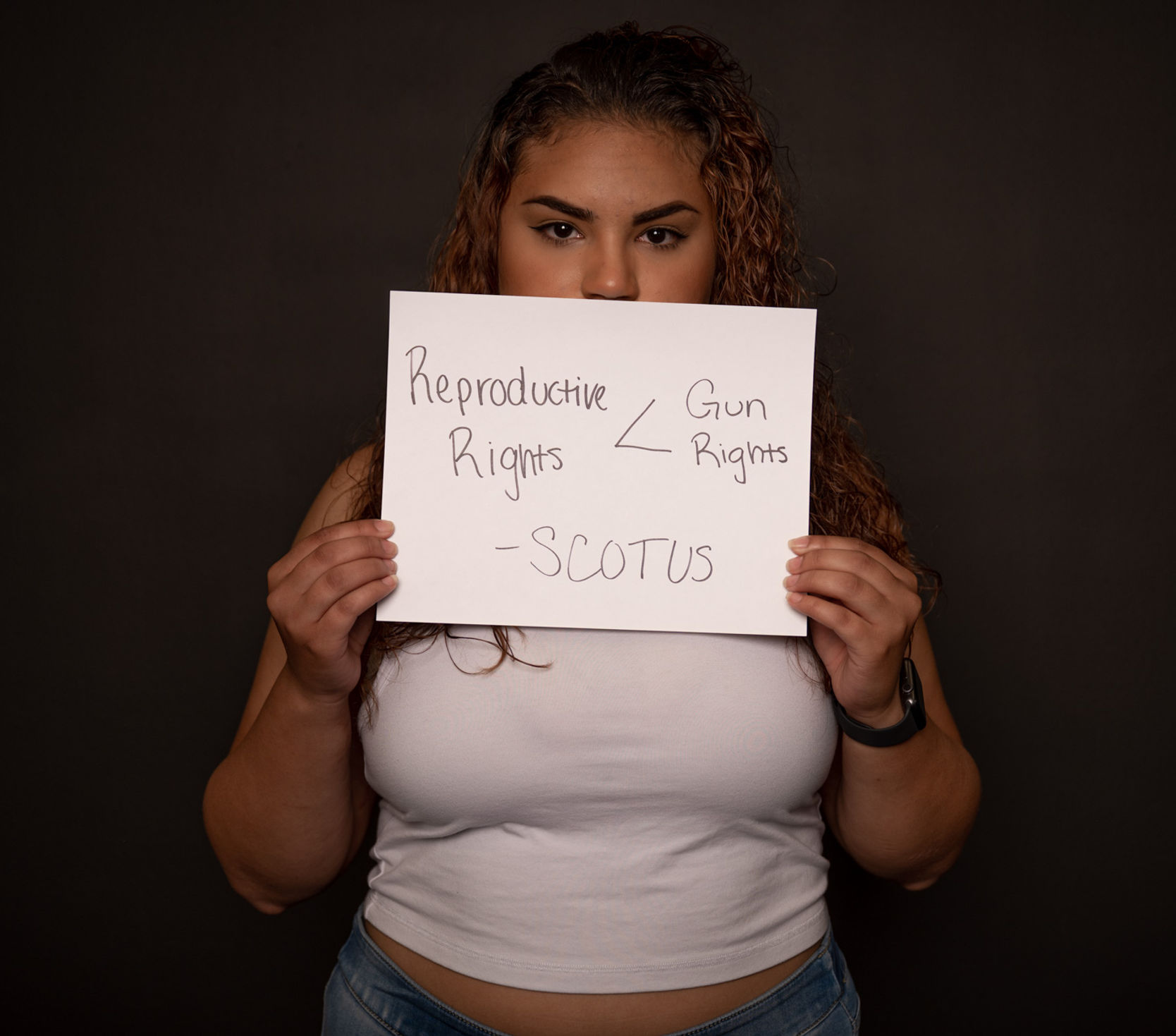 Nayeliz holding a sign partially covering her face that says "Reproductive Rights (less than sign) Gun Rights

- SCOTUS" 