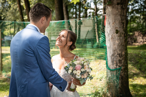 Braut und Bräutigam stehen sich gegenüber, Braut hat Brautstrauß, Hintergrund mit Bäumen