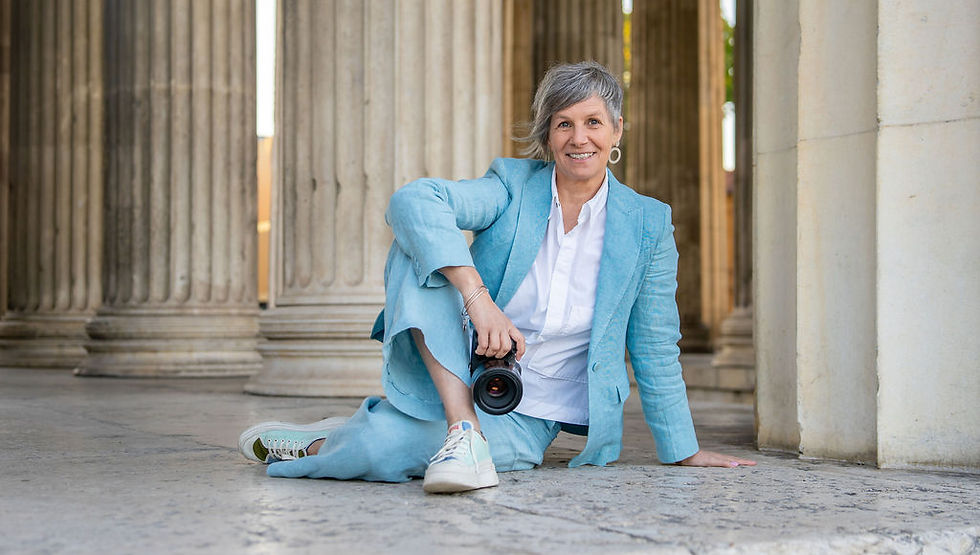 Frau lächelt, hat eine Kamera in der Hand, sitzt auf dem Boden, rechtes Bein über das linke Bein geschlagen, im Hintergrund Säulen