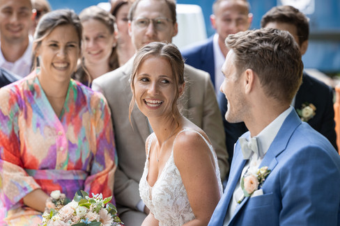 Braut lacht Bräutgam an, beide sitzen auf Stuhl