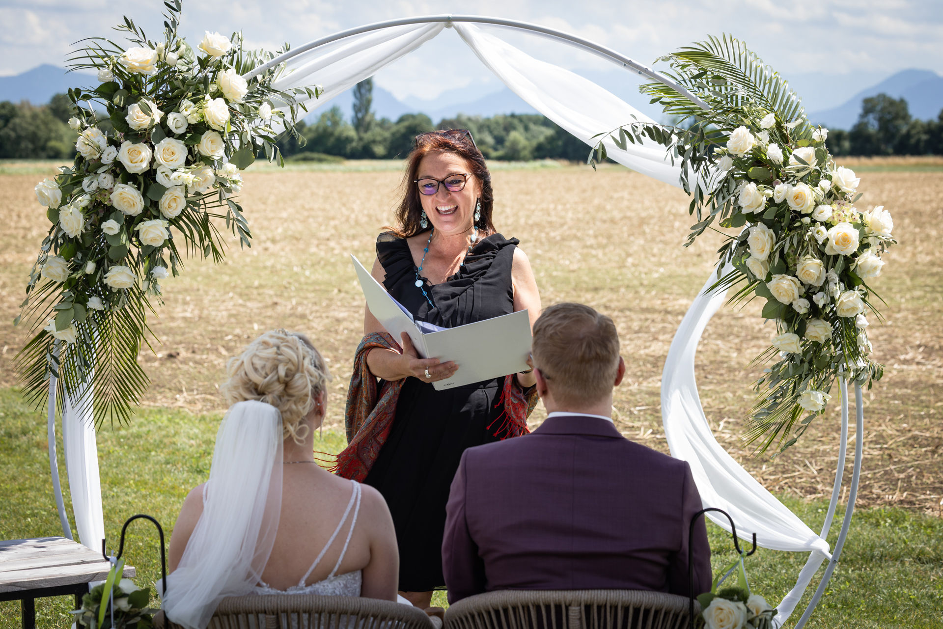 Hochzeit mit freier Trauung draußen im Tropenhaus Weihenlinden in Oberbayern, Traurednerin mit Buch unter Traubogen, Brautpaar sitzt davor