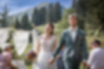 Braut und Bräutigam gehen Hand in Hand, im Hintergrund Berge, Gäste und Traubogen