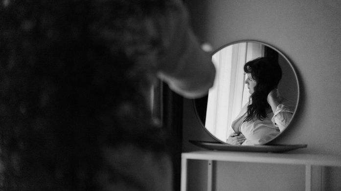 Frau betrachtet sich im Spiegel, Selbstliebe, im Zimmer mit kleinem Tisch.