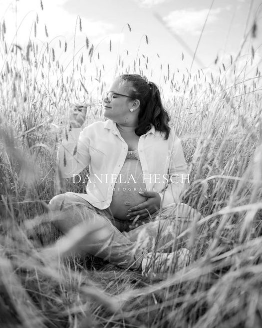 Schwangere im Weizenfeld, Bluse ist geöffnet und zeigt ihren Babybauch, sie shcnuppert an einem Blümchen