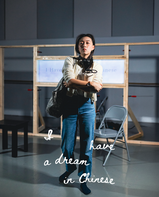 A young actor is rehearsing in front of a wooden-framed backdrop, holding a portable travel water bottle and a travel bag.