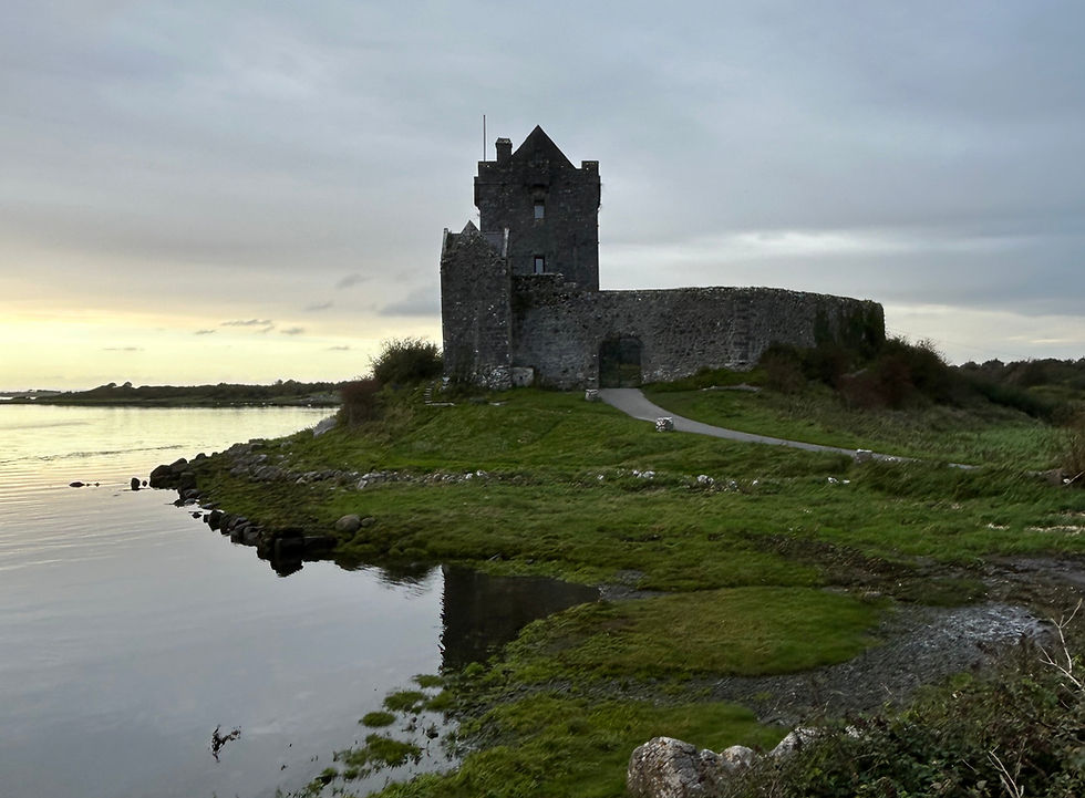 Stone castle by a calm lake at sunset, surrounded by green grass and clouds. Serene and historical mood with a paved path leading to the castle.