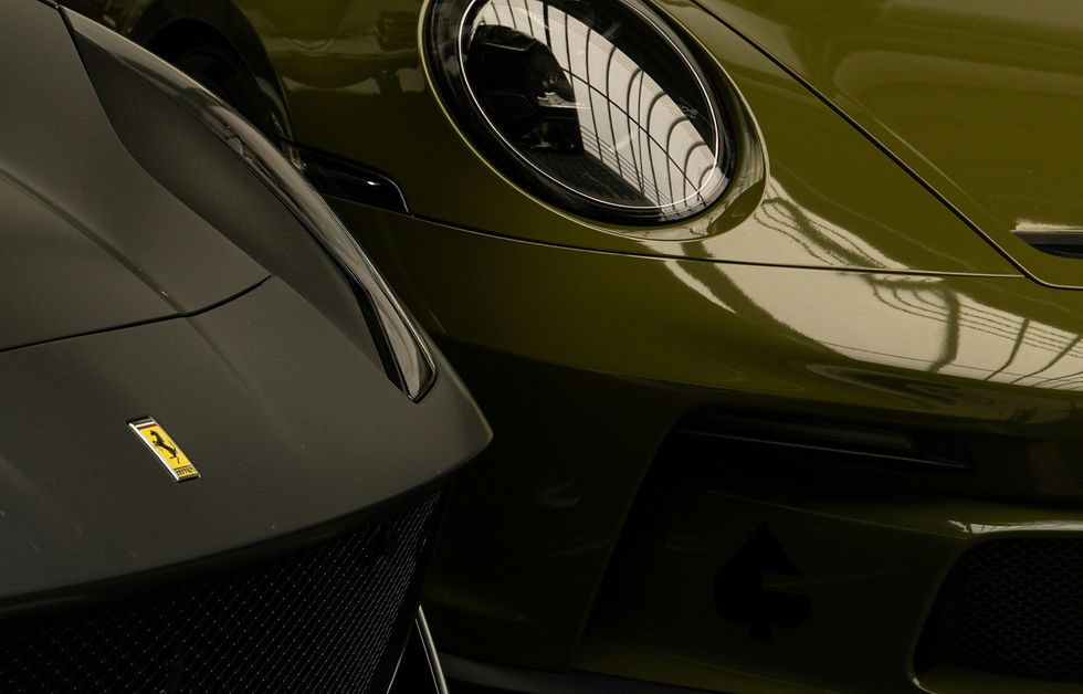 Close-up of two Ferrari sports cars nose to nose, matte black and olive green, with glossy reflections and a Ferrari badge.