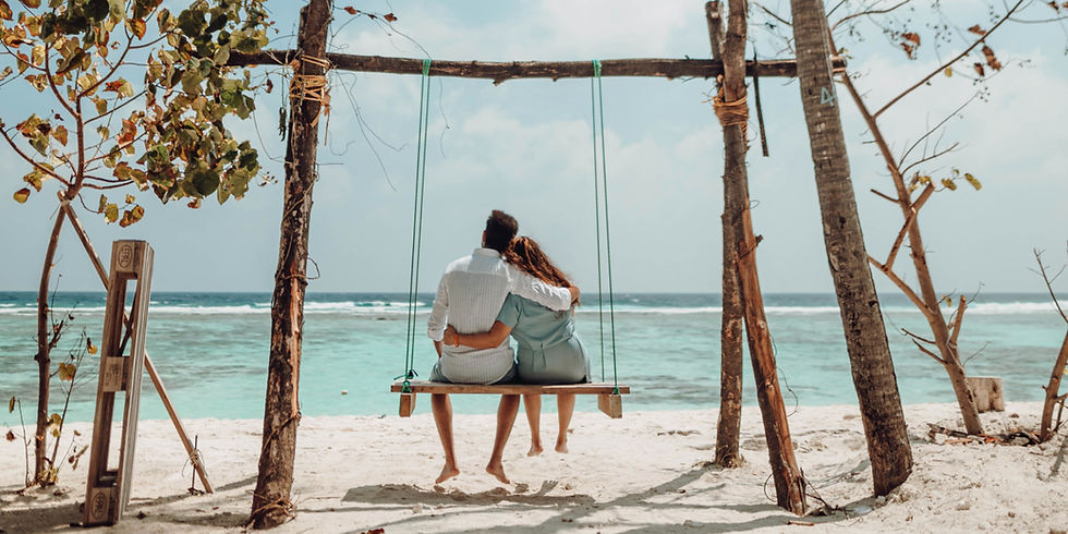 Couple on a swing embraces by the beach, facing the turquoise sea under a clear blue sky. Trees frame the scene, evoking a serene mood.