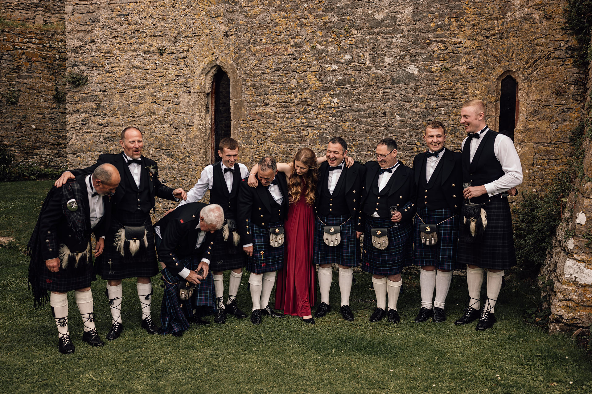 Groomsmen dressed in kilts posing together for a photo in front of a castle wall. There is one bridesmaid in a long red dress in the middle of them all and one of the gentleman's kilts is falling down.