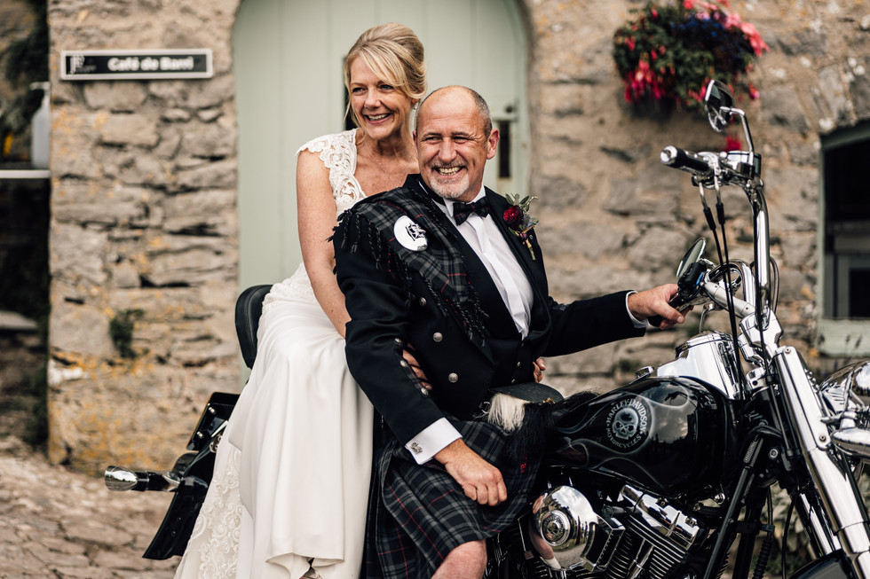Bride and groom sitting the back of a Harley Davidson motorbike.