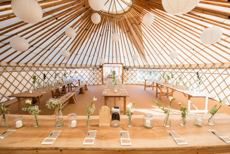 Inside a wedding yurt, there are long wooden rustic tables dressed wth white and green flowers in glass bottles.