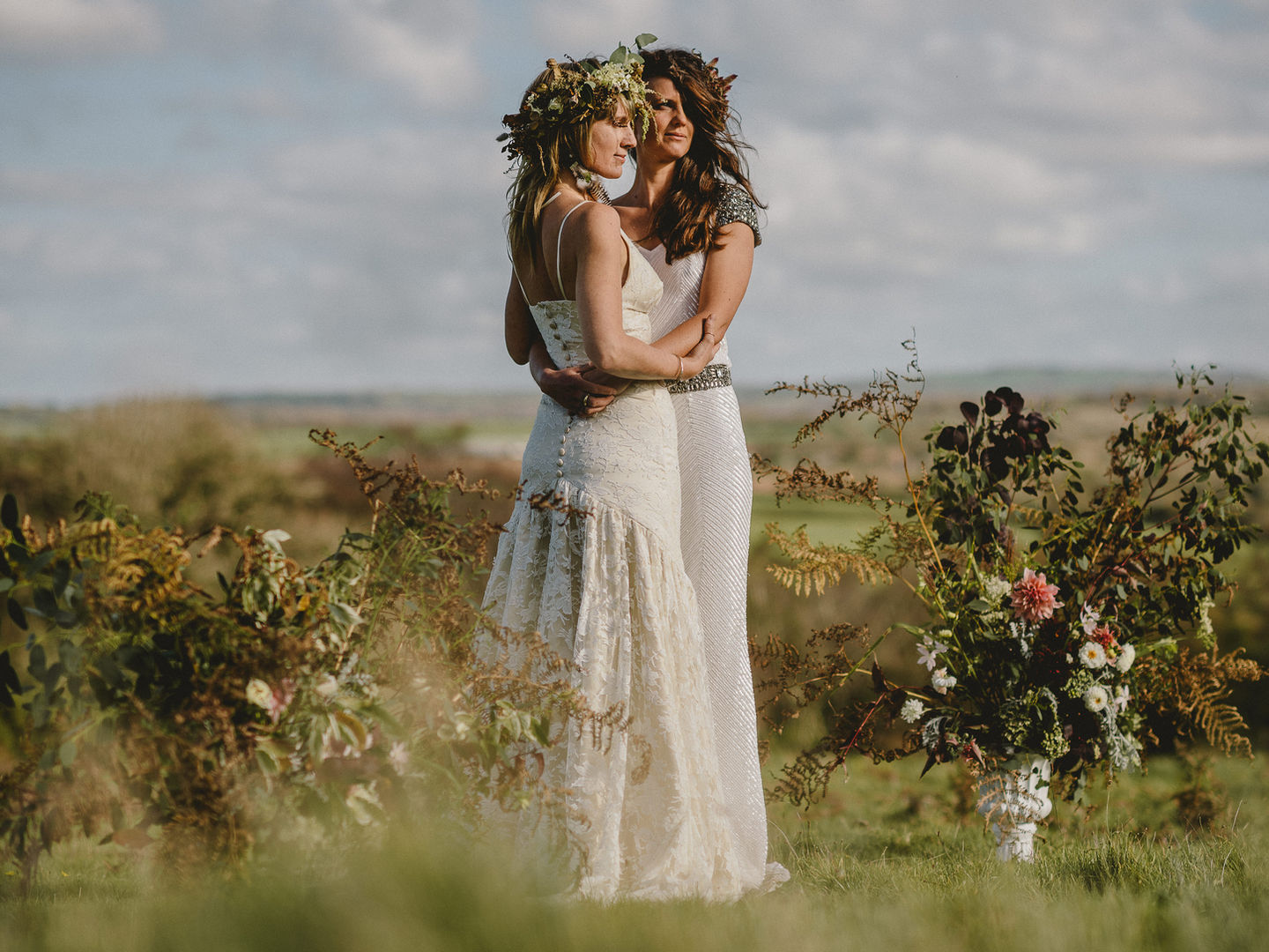 Couple shot of two brides embracing in a field and surrounded by flowers.