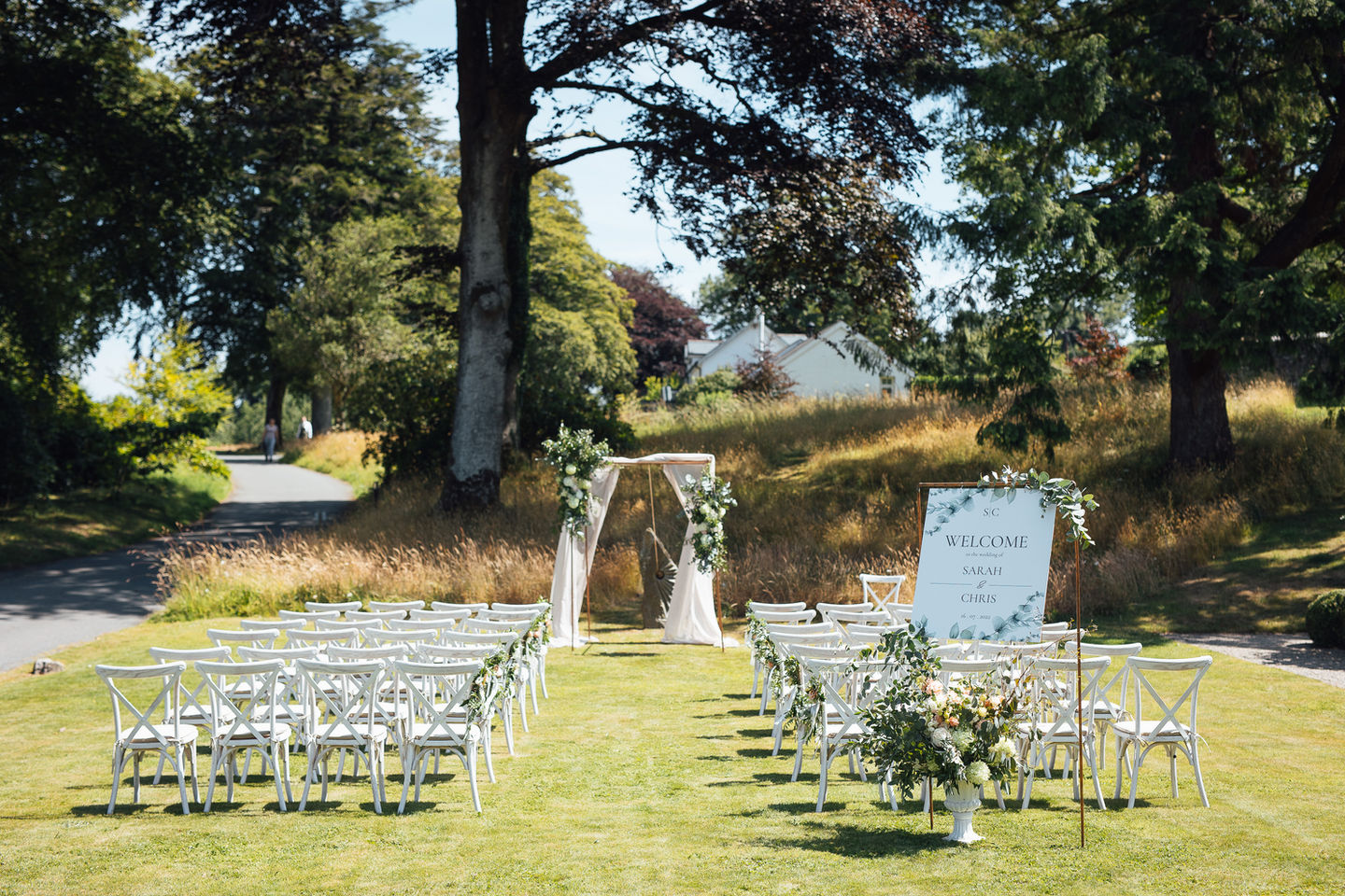 Outdoor wedding ceremony on a lawn with tree's in the background. There are white cross back chairs and a copper arch decorated with cream fabric and large floral displays.