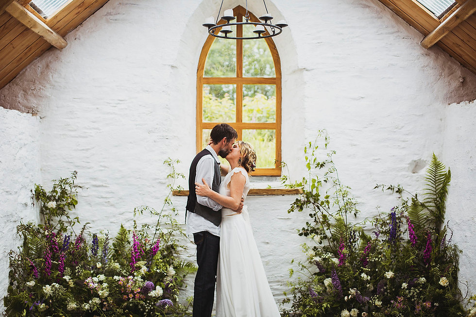 Bride and groom kissing inside a whitewashed church. There is an arched wooden window and wildflower displays behind them.