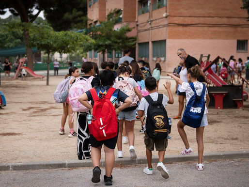 Children with backpacks gather and walk into a school playground beside a brick building, suggesting the start of class.