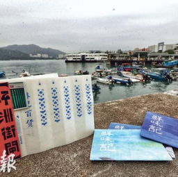 Product photograph showing a book titled 'Peng Chau Street Gates' in Chinese propped up on a jetty edge with a seaview and parked fishing boats in the background.