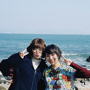 Color photo of SeoiSeoiNim, two young women posing together on a rocky shoreline against a blue ocean and hazy mountains; the woman on the left wears glasses, a navy zip-up jacket, and makes a peace sign, while the woman on the right wears a colorful floral dress with red frog buttons and a red mushroom crossbody bag, also making a peace sign.