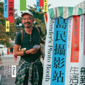 Color photo of Chun, a smiling man with a mustache and beard wearing a dark t-shirt, a baseball cap, and a plaid shirt tied around his waist, standing next to a vintage cart with a large 'Islander's Photo Booth' banner at the Inter-Island Festival, with colorful location signs and text in the background.