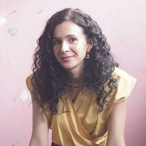 Color photo of Iris and Louisa, two smiling women standing behind a dark table; Iris on the left has straight dark hair and wears a white t-shirt and black pants, while Louisa on the right has long curly dark hair and wears a yellow draped top, both leaning forward against a distressed pink wall with peeling paint.