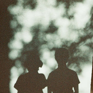 Artistic photo titled inlightofliving, showing the dark silhouettes of two people standing against a textured wall, backlit by soft, blurry, white light filtering through out-of-focus leaves or clouds.