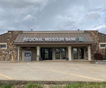 Exterior view of a one-story stone and metal building with the sign "REGIONAL MISSOURI BANK" above the entrance, featuring large front windows, an ATM, and a mostly empty concrete parking lot under a cloudy sky.