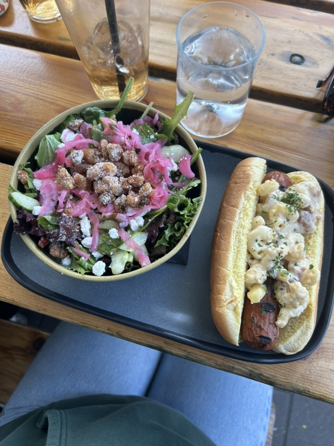 Mixed greens salad with pickled onions, candied pecans, and crumbled goat cheese, alongside a mac and cheese hot dog on a wooden table.