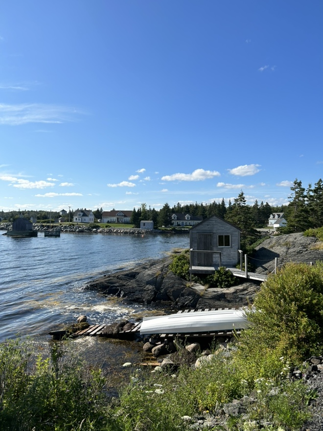 Coastal view of a small harbor town with a white boat on a wooden dock, rocky shoreline, and a light gray boathouse.