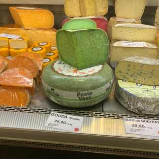 Assortment of cheeses, including Gouda Basilic (green pesto flavored), displayed in a refrigerated case with price tags.