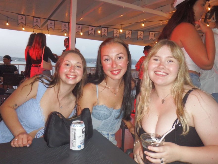 Three young women, smiling and enjoying drinks outdoors on a boat.