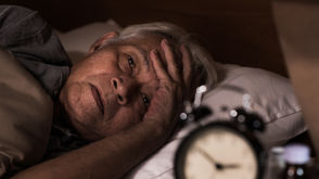 Man struggling to fall asleep in bed, looking troubled, with an alarm clock beside him