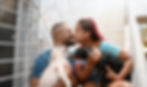 A young couple stands outside, holding their respective small dogs in their arms, leaning forward to share a kiss, displaying a heartwarming moment of affection between the couple, accompanied by their beloved pets