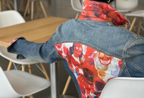 A woman sits in a cafe wearing a handcrafted Go Coco! jean jacket with an Alexander McQueen scarf sewn on it