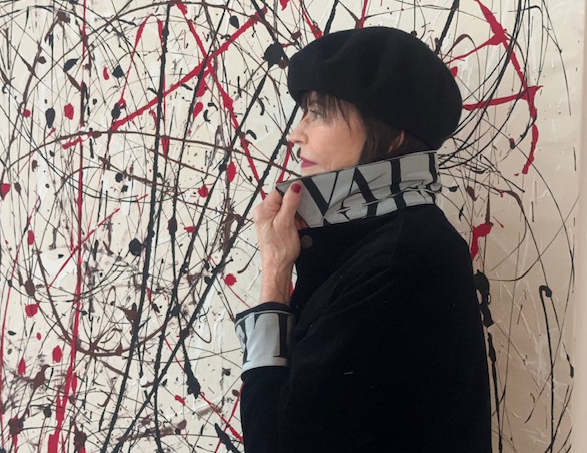 A woman is beside a wall spattered with paint wearing a designer jean jacket with a white and black Valentino scarf