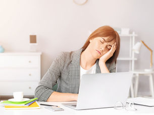 Woman in checked blazer asleep at a desk with a laptop. Books, cup, pens nearby. Bright, minimal office setting suggests fatigue.