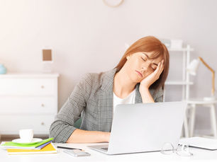 Woman in checked blazer asleep at a desk with a laptop. Books, cup, pens nearby. Bright, minimal office setting suggests fatigue.