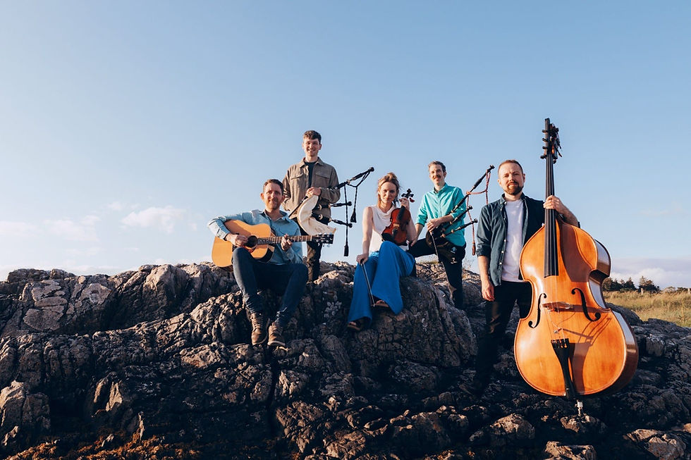 Breabach pose on a rocky shore under a blue sky, holding guitar, violin, bagpipes and double bass, smiling.
