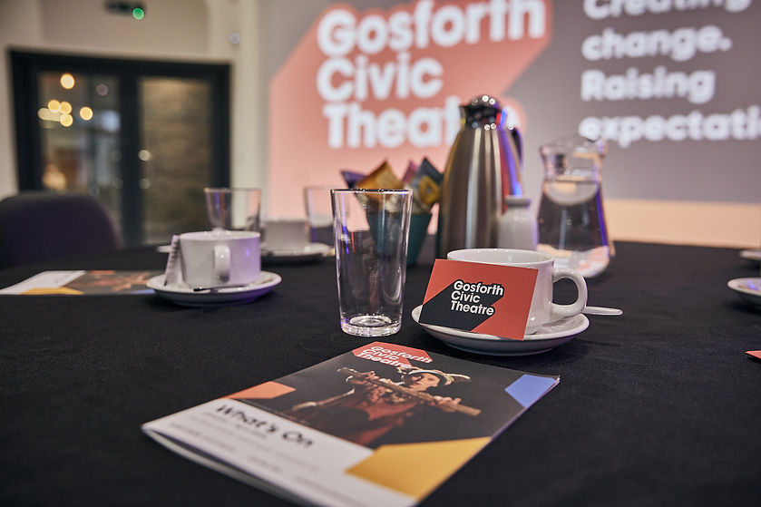 Gosforth Civic Theatre business cards placed on mugs with glasses, water and What's On Guides on a table with a black table cloth. Blurry background showing a projected image on the wall.