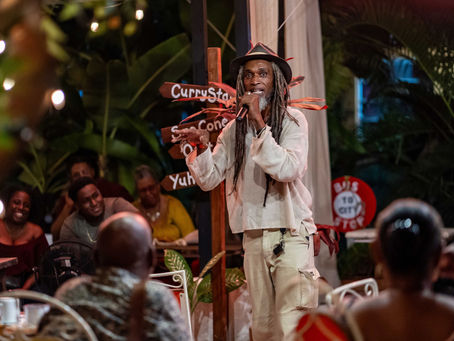 Bajan man storyteller standing surrounded by seated smiling listeners. Outside at night in a garden