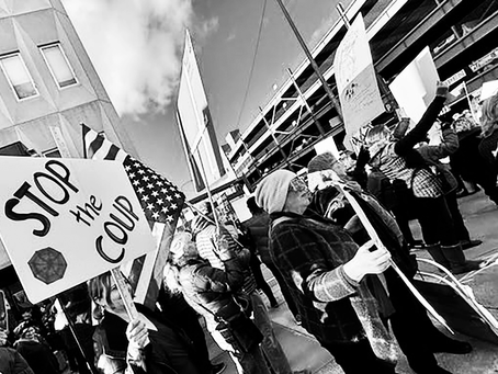 Black and white image of protest rally against Trump with a "Stop the Coup" sign prominent.