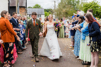 Newlyweds walk through confetti as guests cheer, A glorious Farm Wedding in Aldbourne, Wiltshire.