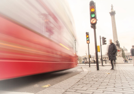 A long-exposure photograph at Trafalgar Square, London, showing a blurred red double-decker bus rushing past a pedestrian near traffic lights. Nelson’s Column and the National Gallery are visible in the background. Captured from a low perspective by Janusz Miarka.