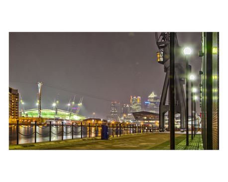 A night-time photograph of Emirates Royal Docks featuring historic dock cranes, glowing street lamps, and the illuminated O2 Arena against the modern skyline of Canary Wharf, captured by Janusz Miarka.