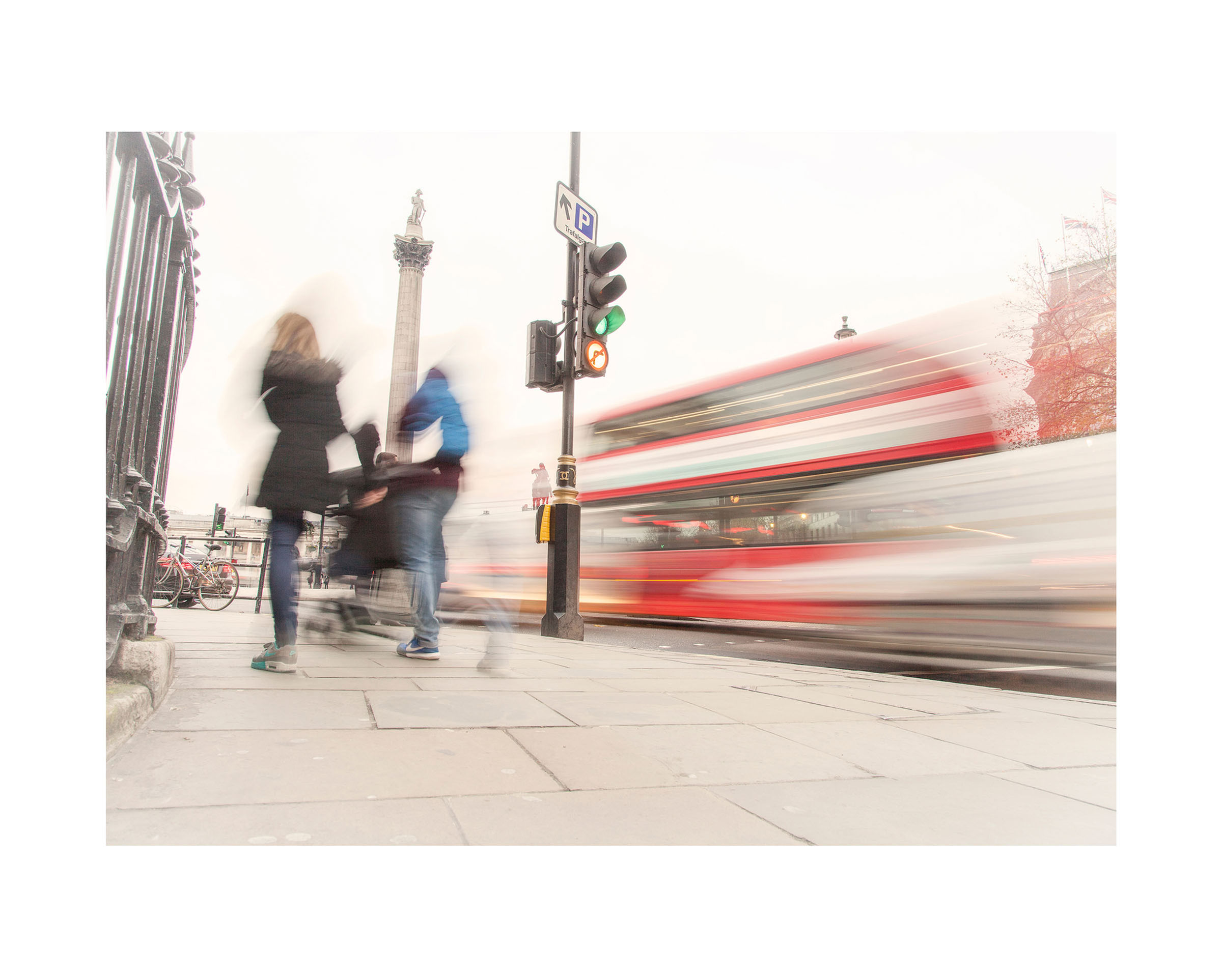 People walking near a London traffic light, with a red double-decker bus passing by and Nelson's Column in the background.