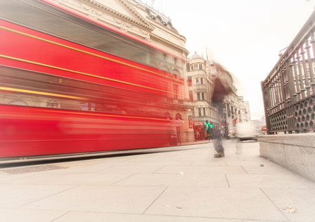 A long-exposure photograph of a red double-decker bus speeding past historic buildings in London's theatre district, with a blurred pedestrian crossing the street. Shot from a low perspective with a tripod by Janusz Miarka, part of the Motion in London fine-art series.