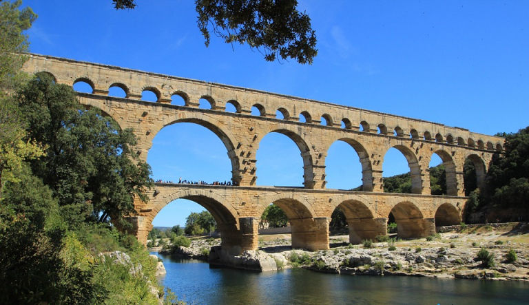The Pont du Gard Roman aqueduct spanning a calm river, reflected in the water and surrounded by trees.