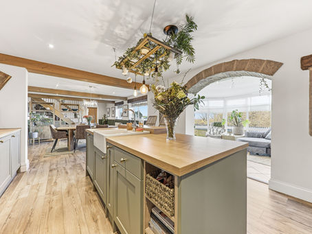 Spacious kitchen with wooden floors, a central island, and hanging plants. Cozy seating and large windows in the background create a warm ambiance.