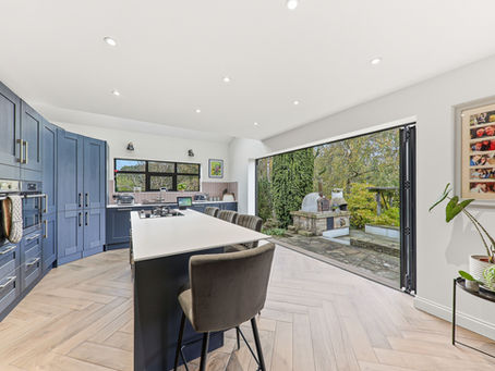 Modern kitchen with blue cabinets, central island, and garden view. Open patio doors, herringbone floor, and hanging family photos.